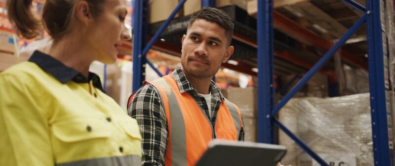Two warehouse team members in safety vests discussing logistics in front of pallet racks