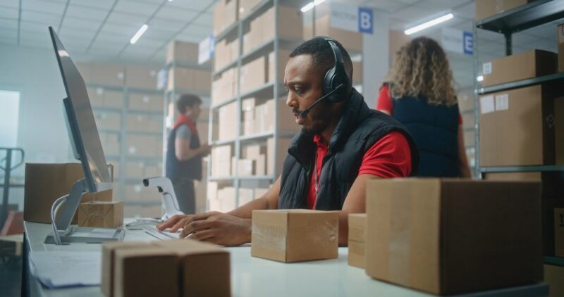 Warehouse worker wearing a headset managing orders on a computer surrounded by packages