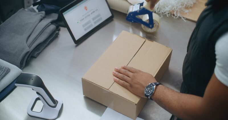 Hands preparing and sealing a cardboard box on a work table using a tape dispenser