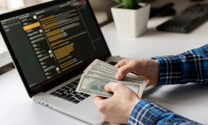 A person in a plaid shirt counts dollar bills at a desk. A laptop displaying code is open nearby, suggesting coding work