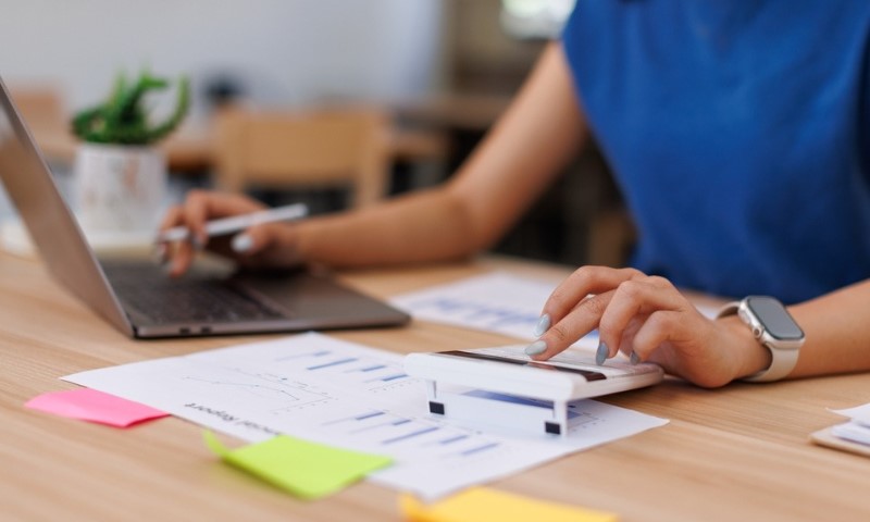 A person in a blue shirt, using a calculator with one hand, and typing on a laptop with the other