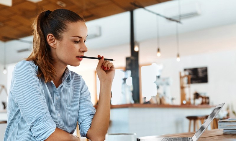 A woman in a blue striped shirt is deep in thought, holding a pen to her lips, while looking at a laptop